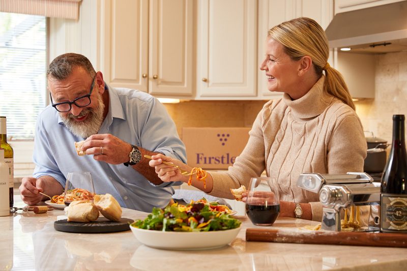 A couple enjoys a meal together in a kitchen, sharing pasta and salad with wine. The man has a beard, and the woman has her hair tied back. A pasta machine and a Firstleaf box are on the counter. The atmosphere is warm and casual.