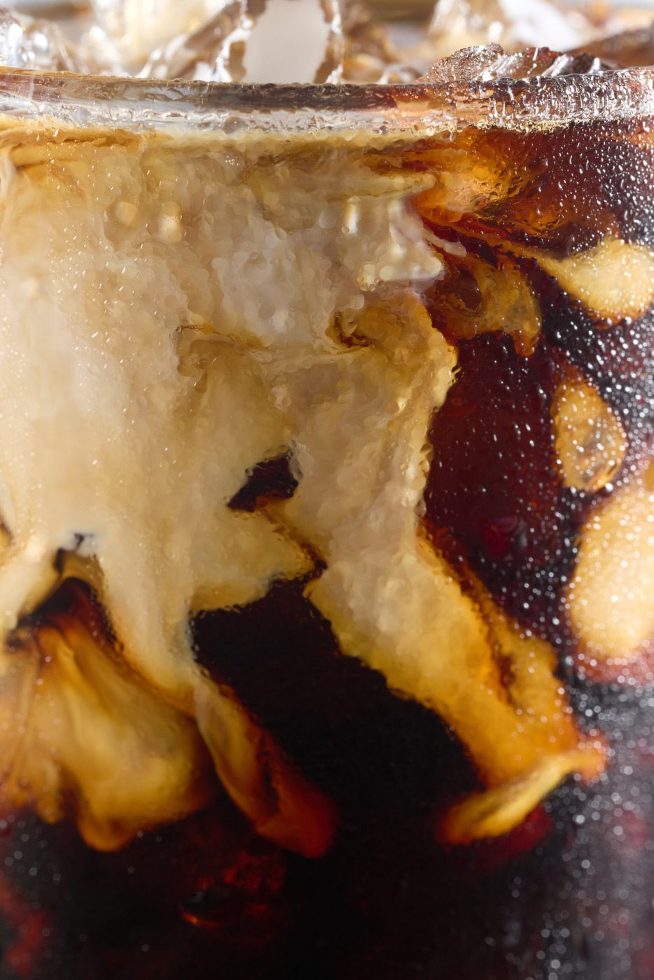 Close-up of a glass of iced coffee with cream swirling through dark coffee, creating a marbled pattern. The surface shows ice cubes and a cold, refreshing texture.