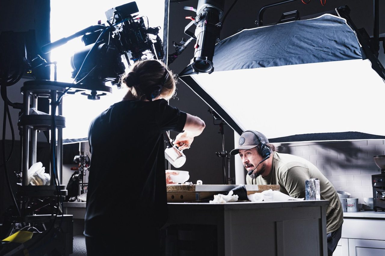 A woman wearing headphones pours liquid while a man observes closely on a film set. They are surrounded by professional lighting and camera equipment, focused on capturing a scene in a kitchen setting.