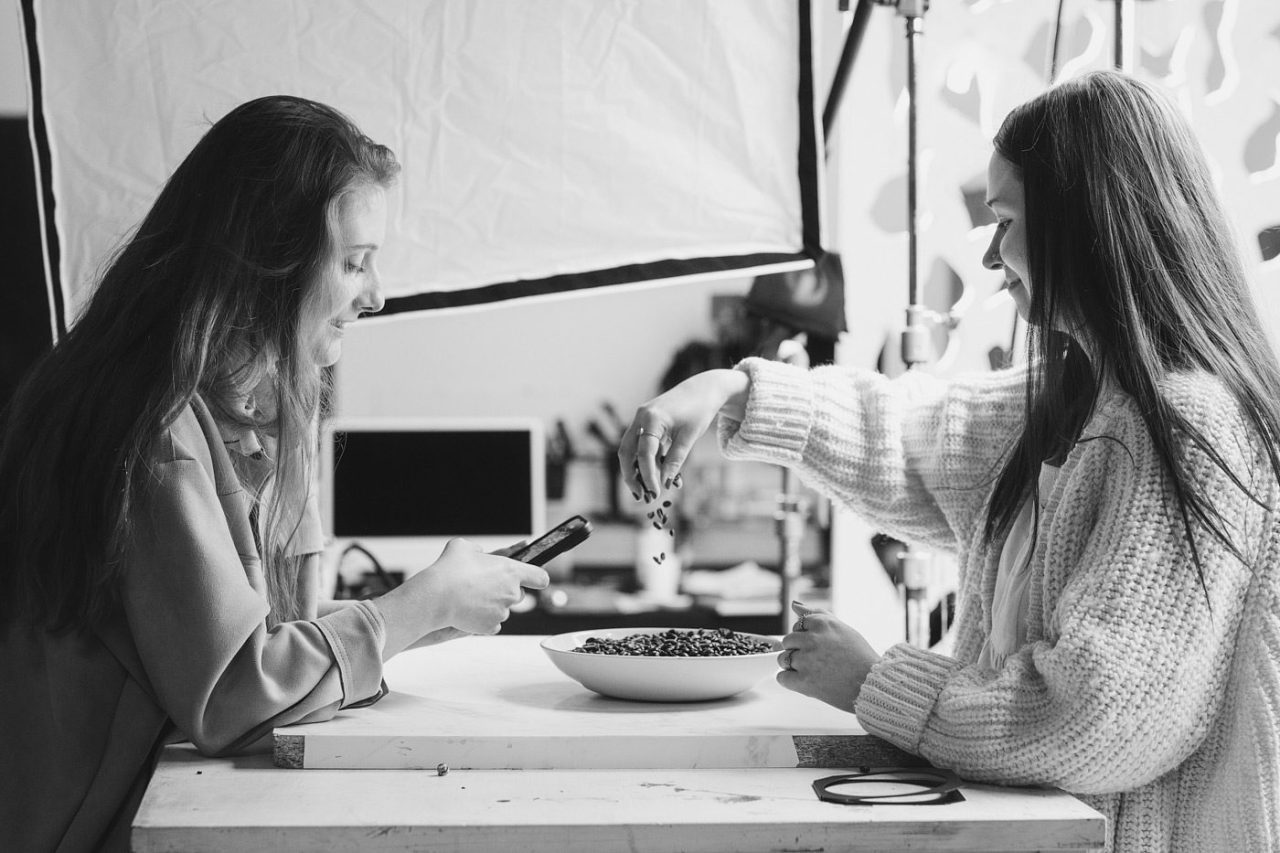 Two women in a studio setting, smiling and engaged in a food photography session. One holds a smartphone to capture the shot, while the other adds berries to a bowl. Professional lighting equipment is visible in the background. Black and white image.