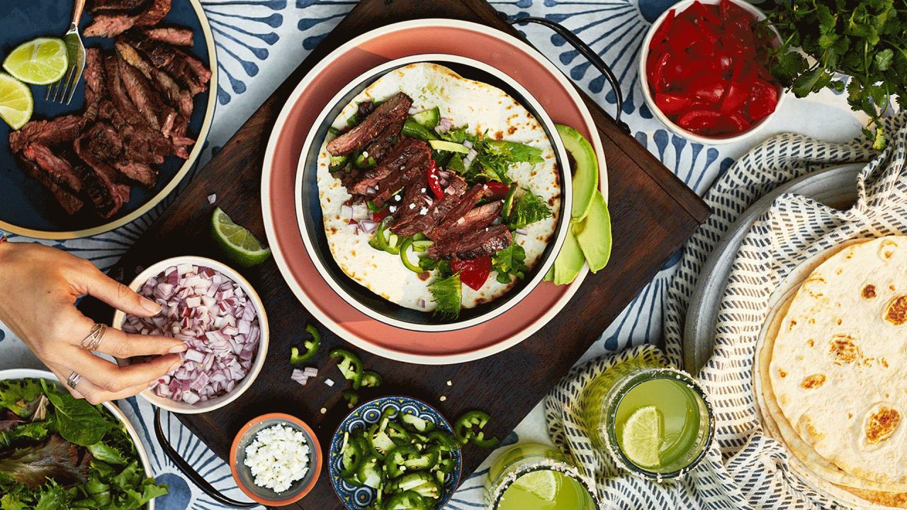 A colorful spread of taco ingredients showcases grilled steak on a tortilla, sliced jalapeños, chopped onions, lime wedges, diced tomatoes, and fresh cilantro. Capturing the essence of food photography for restaurants, two glasses of green beverages and a hand reaching for onions complete the scene.