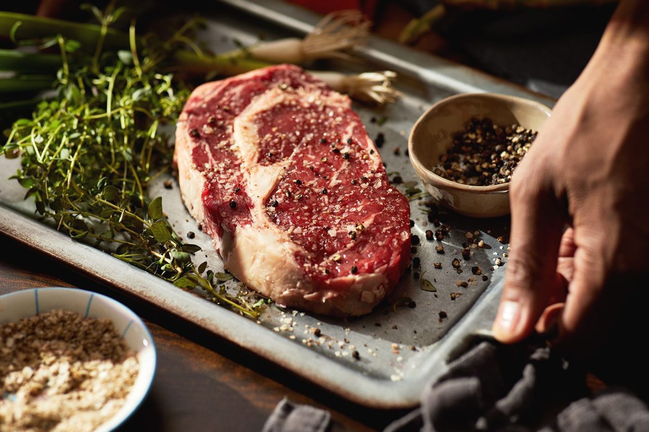 A hand holds a cloth near a raw ribeye steak on a tray, sprinkled with coarse salt and pepper. Fresh herbs, a small bowl of peppercorns, and sliced green onions are arranged nearby, capturing the essence of rustic food photography.