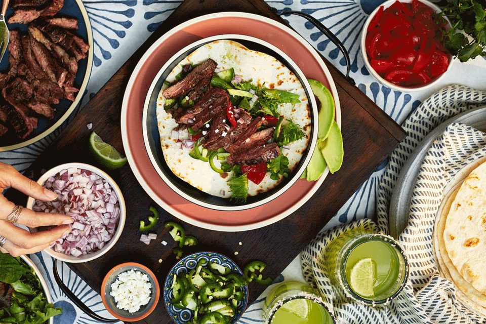 A vibrant food photography setup showcases a taco on a plate filled with grilled meat, cilantro, and lime. Surrounding the taco are small bowls of chopped onions, jalapeños, and cheese. A hand reaches for the onion bowl. A refreshing drink and lime wedges complete this captivating scene.