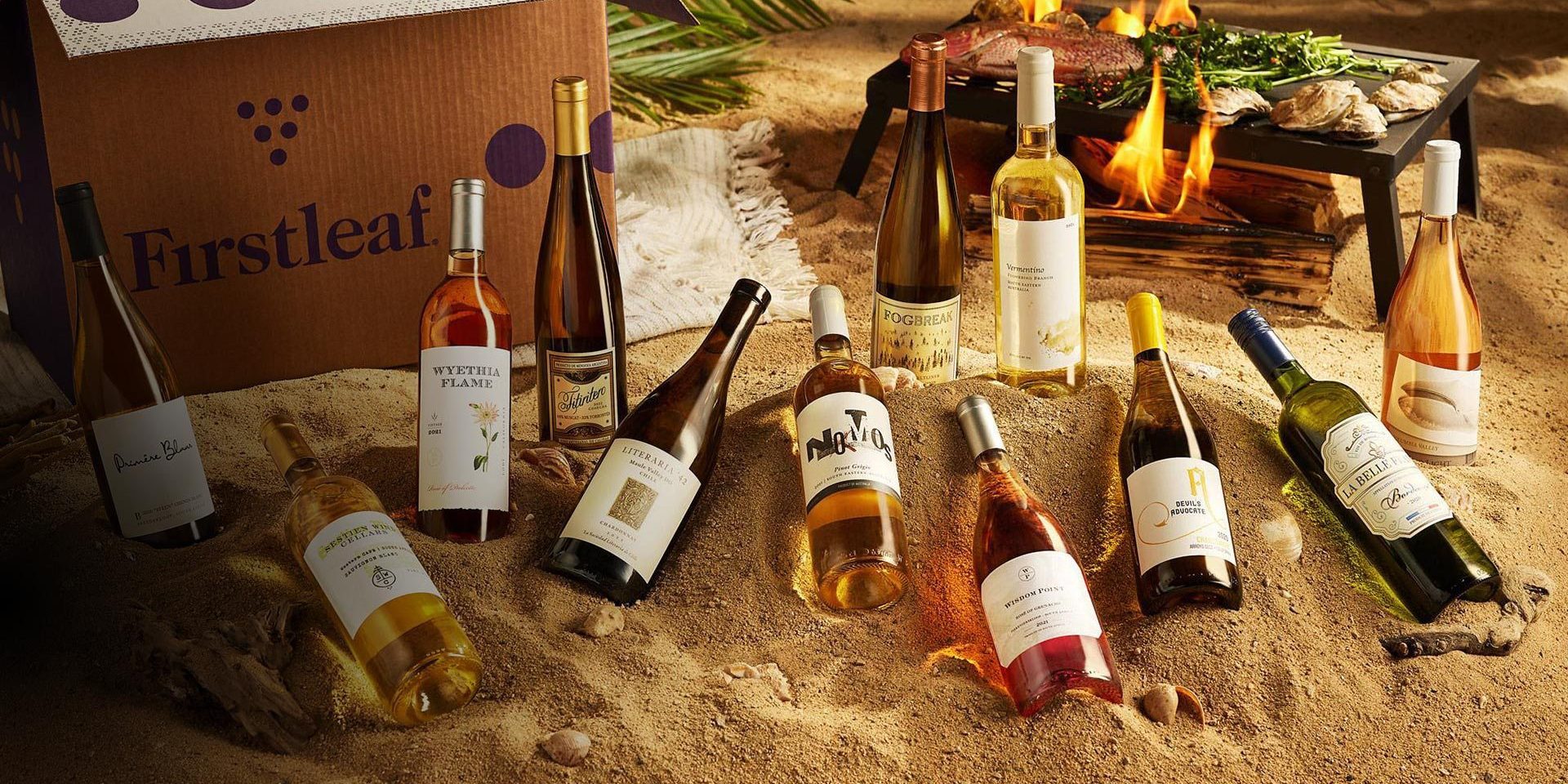 A variety of wine bottles arranged on a sandy beach setting with a Firstleaf box in the background. A small grill with vegetables and oysters is nearby. Lush green foliage is partially visible.