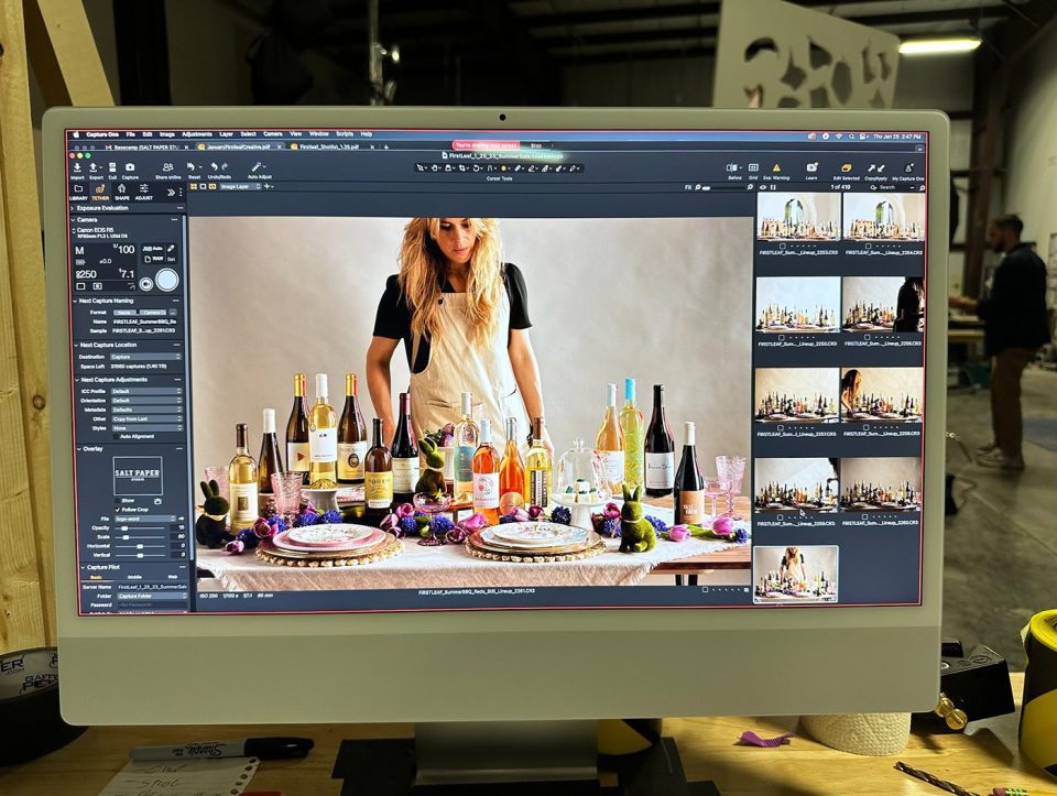 A computer screen displaying photo editing software shows a woman arranging a table with various bottles, fruits, and tableware. Several thumbnail previews of similar images are visible on the right side of the screen.