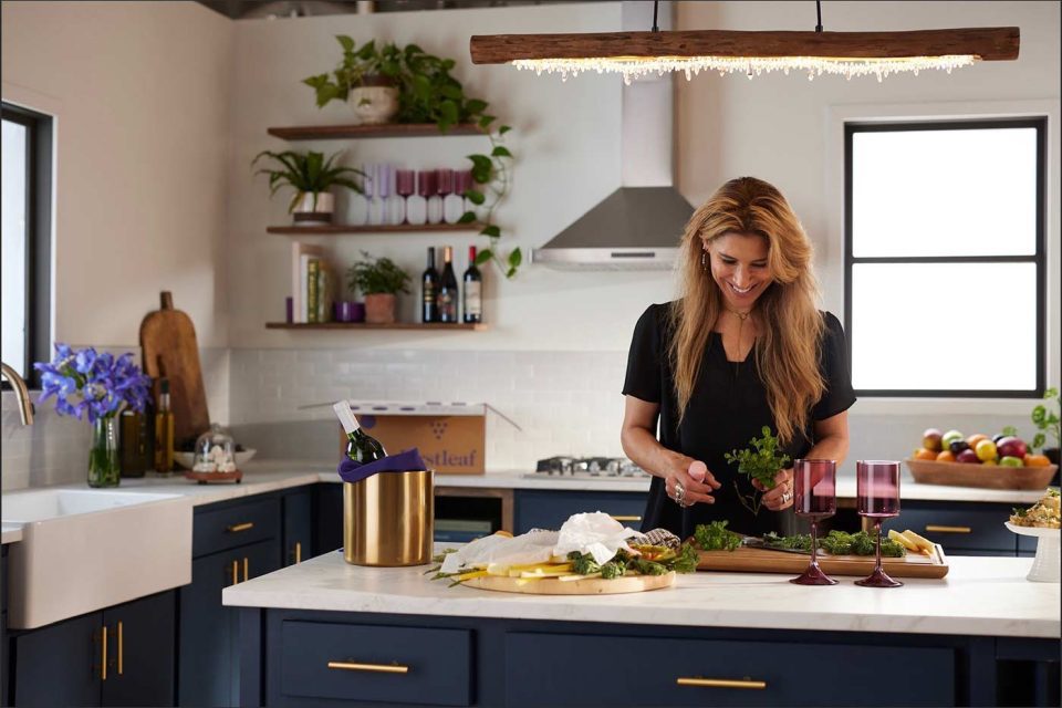 A woman in a modern kitchen chops herbs on a wooden board. The countertop features wine glasses, a bottle in a cooler, and a tray of cheese. Shelves hold plants and wine bottles, and there are fruits in a bowl nearby.