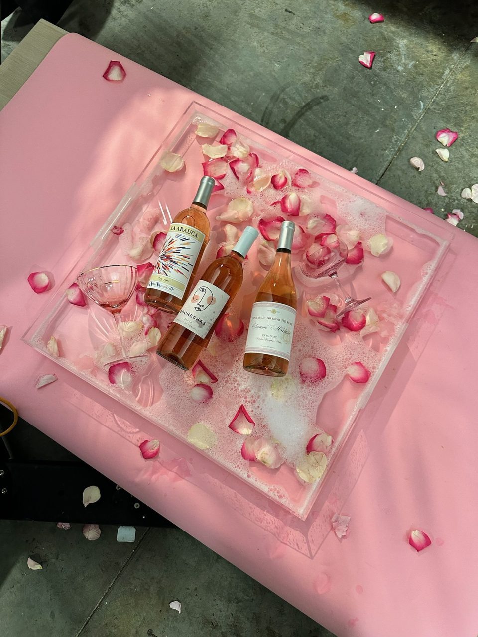 Three bottles of rosé wine on a pink surface, surrounded by scattered rose petals and a wine glass. The setting has a romantic and elegant feel with a mix of colors and textures, including pink foam.