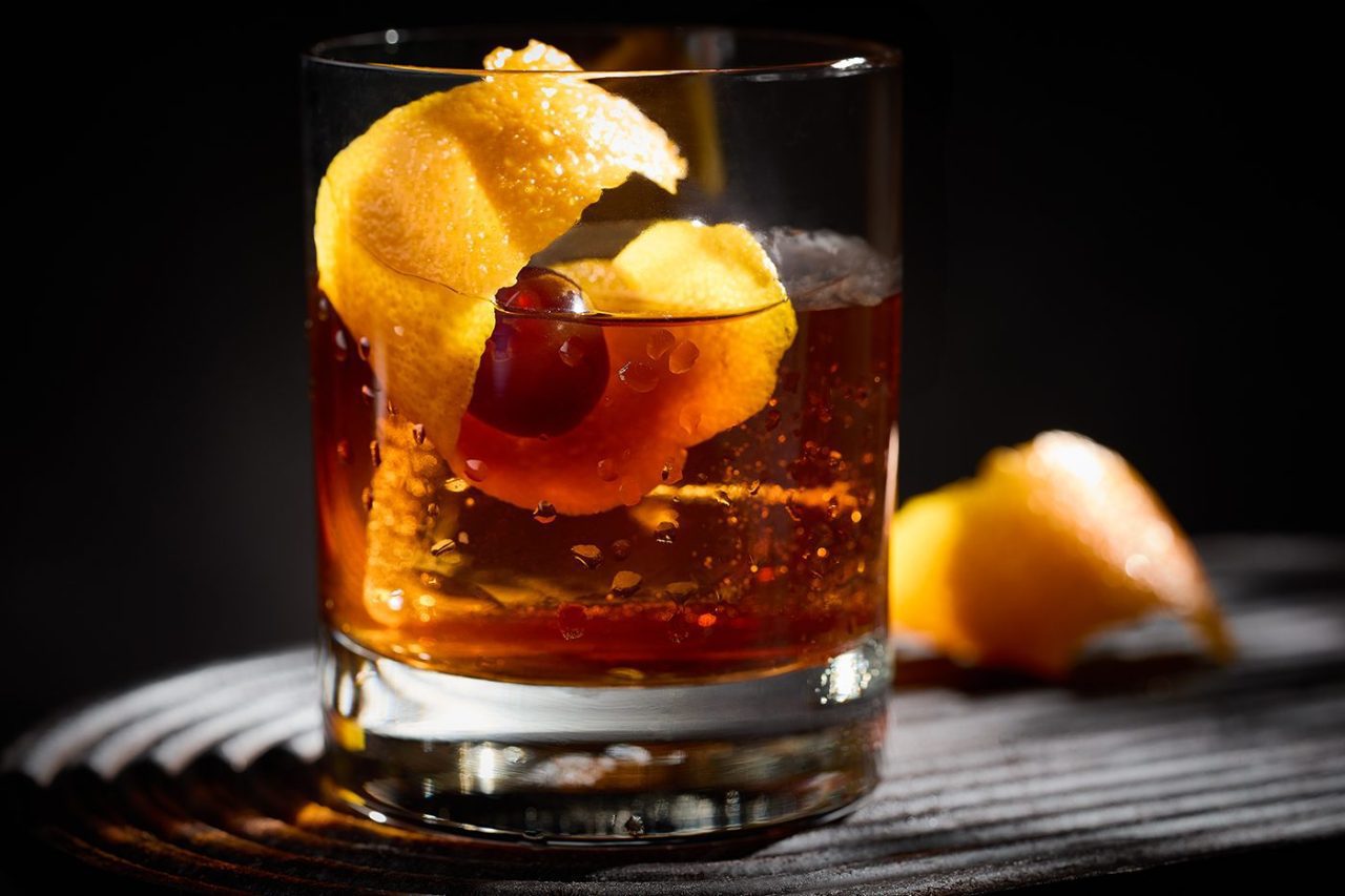 A clear glass holds a dark-colored cocktail garnished with a twist of orange peel and a cherry. The drink is set against a dark background, with a piece of orange peel resting nearby. The lighting creates a warm, inviting glow.