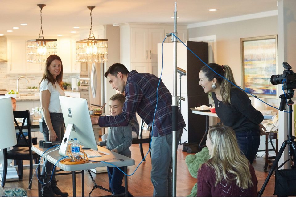 A group of people are gathered around a computer in a well-lit kitchen setting. They are smiling and engaging with the screen. Camera equipment is visible, suggesting a photo or video shoot in progress. The space is modern and bright.