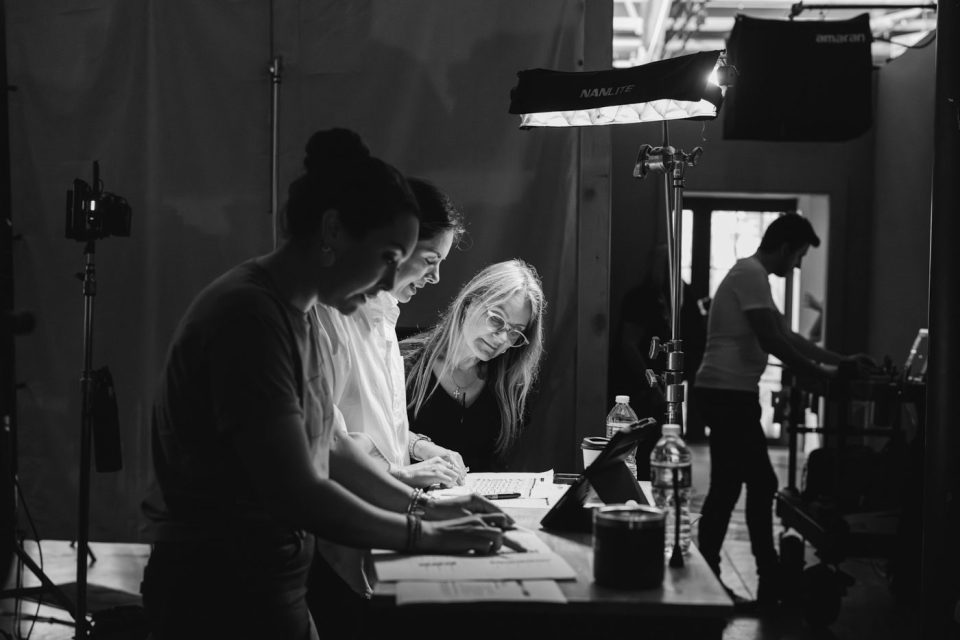 Three people collaborate around a table with papers and a computer in a dimly lit studio. A camera and lighting equipment are visible. One person operates a laptop, while another works on a tablet.