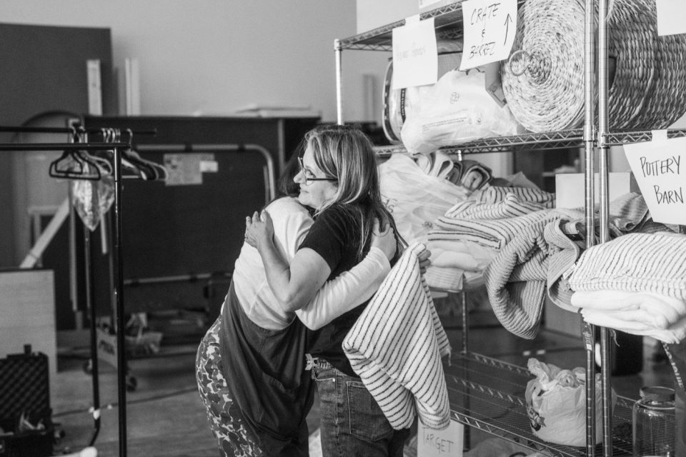 A black and white photo of two people hugging in a room filled with shelves holding blankets and other household items. Signs with labels like "crate & barrel" and "pottery barn" are visible. A clothes rack is partially seen on the left.