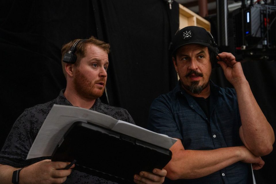 Two men wearing headsets are standing in a dimly lit room. One is holding a clipboard and speaking, while the other listens intently. They appear to be collaborating on a project, with a camera rig visible in the background.