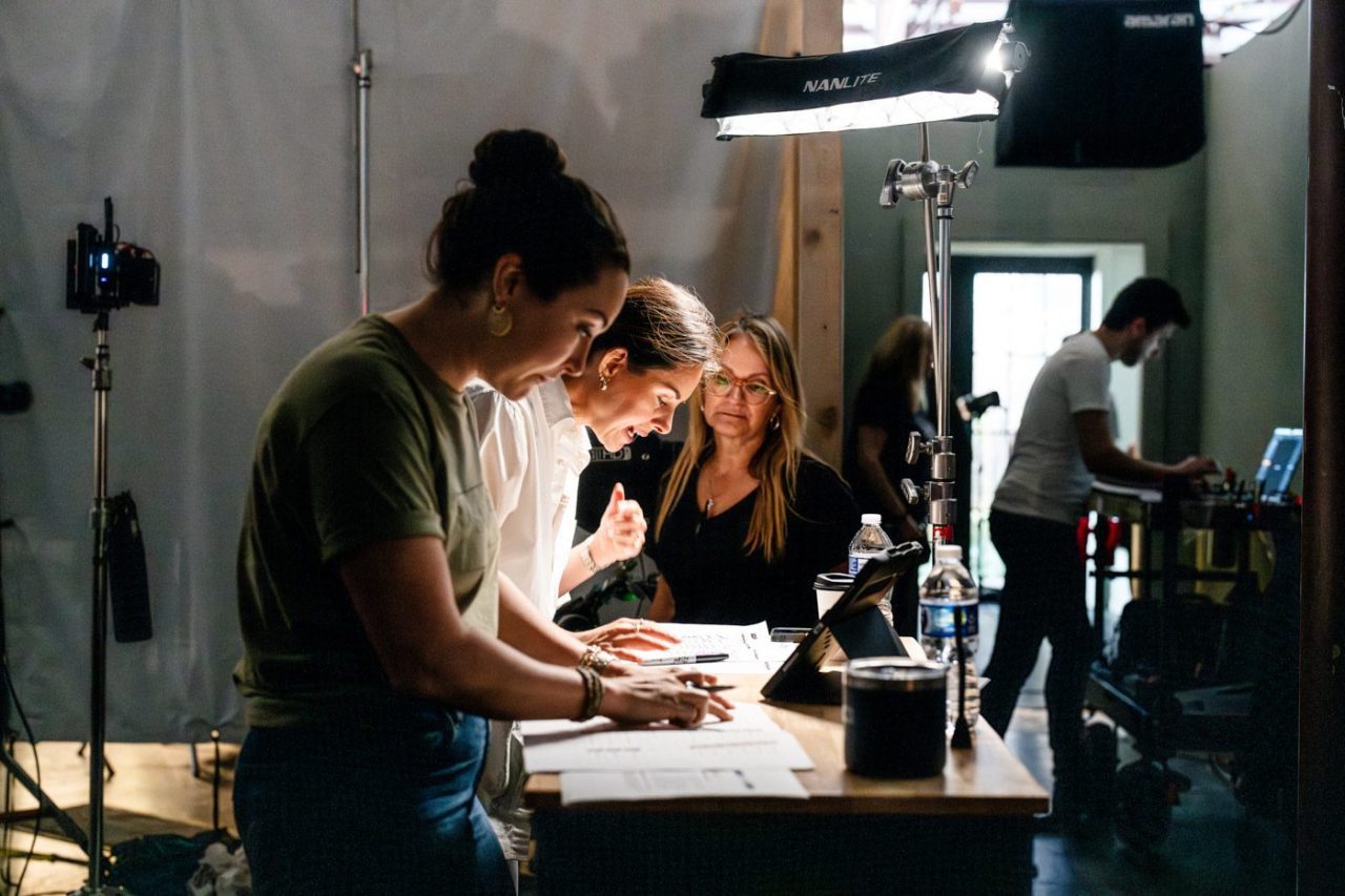 Three people collaborate at a table with papers in a dimly lit room. A woman in the background uses a computer. There is photography equipment around, including lights and a camera, creating a studio environment.