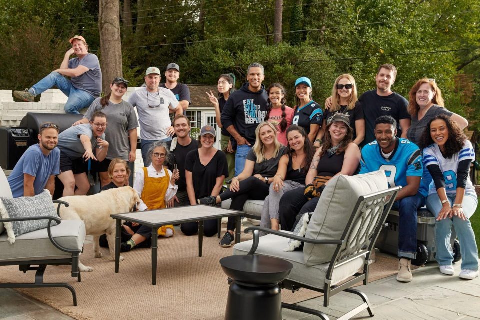 A group of people posing together outdoors on a patio with furniture for a "meet the team" photo. Some are sitting and others standing, all smiling at the camera. A playful dog joins the scene, while trees and a fence provide a charming backdrop in the background.