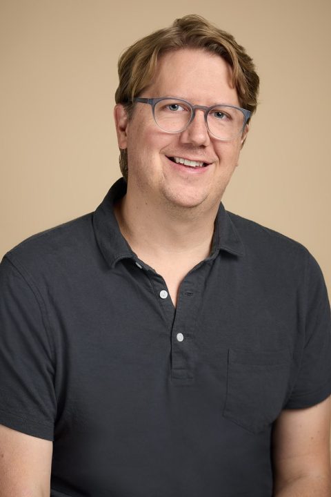 Meet the team: A person with glasses, wearing a dark polo shirt, smiles against a beige background.