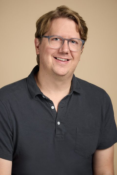 Meet the team: A person with glasses, wearing a dark polo shirt, smiles against a beige background.