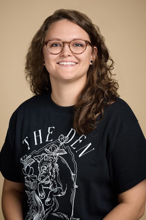 A person with wavy brown hair and glasses smiles warmly at the camera, embodying the spirit of "Meet The Team." They sport a black T-shirt featuring a design and the text "The Den," set against a plain beige background.