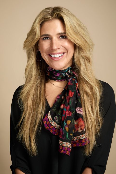 Meet the team: A smiling woman with long blonde hair wears a black blouse and a colorful floral scarf against a neutral beige background.