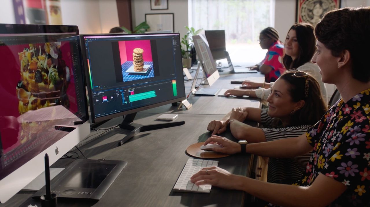 In Our Studio, people are working at computer desks. A large monitor displays a layered cake in video editing software. The room is well-lit, and several individuals are focused on their tasks.