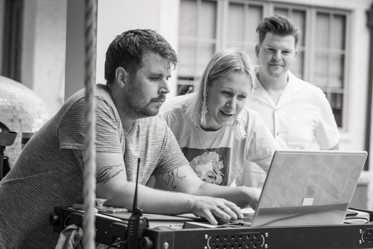 Three people are gathered around a laptop, focused on the screen. One person is typing as another leans in to observe. The third stands behind them, smiling, clearly providing creative direction as they collaborate on this outdoor project.