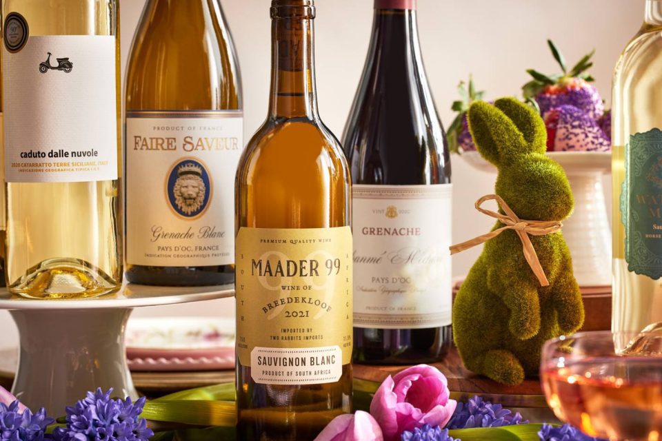 A selection of wine bottles, including Maader 99 Sauvignon Blanc, Caduto Dalle Nuvole, and Faire Saveur, displayed with a green bunny figurine and purple flowers.