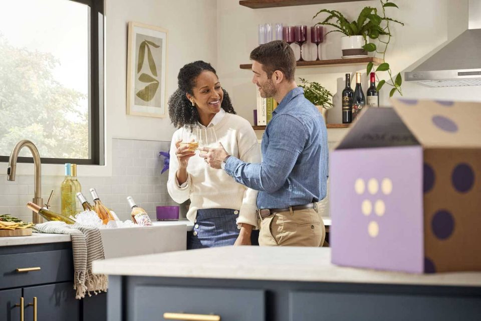 A couple stands in a modern kitchen with navy blue cabinets, holding wine glasses and smiling at each other. A purple box is on the countertop, and shelves with plants and bottles are in the background.