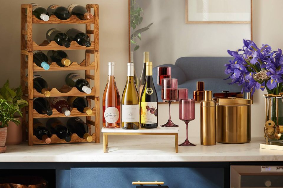 A home bar setup with a wine rack filled with bottles, three wine bottles on a small stand, red wine glasses, golden cocktail shakers, a potted plant, and purple flowers on a marble countertop. A framed picture reflects a blue sofa.