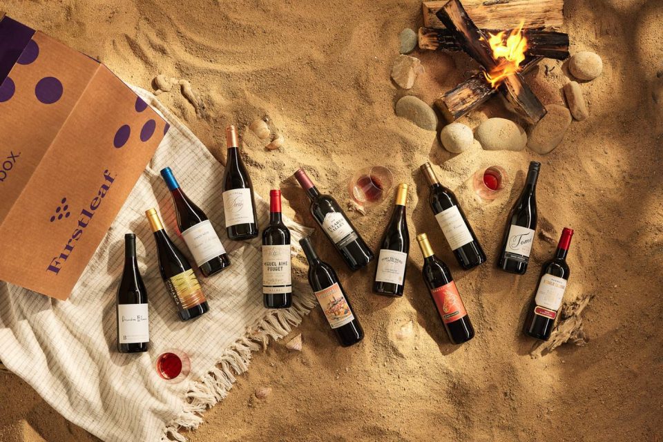 An array of wine bottles is arranged on a sandy beach next to a cozy campfire. A striped blanket and an open box with "Firstleaf" branding are nearby, along with a few poured glasses of wine.