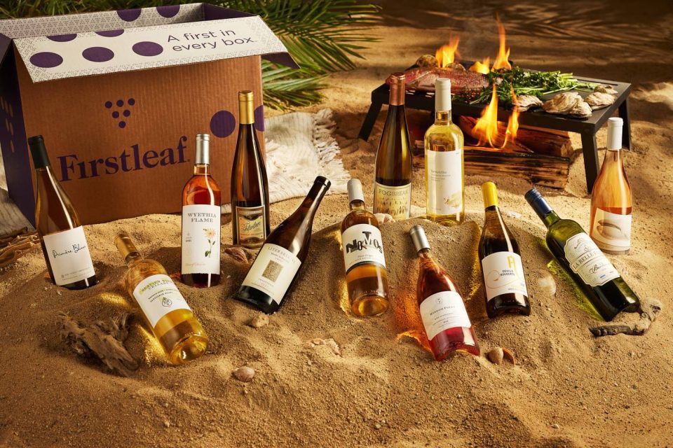 An assortment of wine bottles displayed on sand in front of a small campfire with food cooking. A box labeled "Firstleaf" is on the left, featuring the slogan "A first in every box." 연식을 외부에 명시하지 않은 다양한 와인 병가 모여 있다.