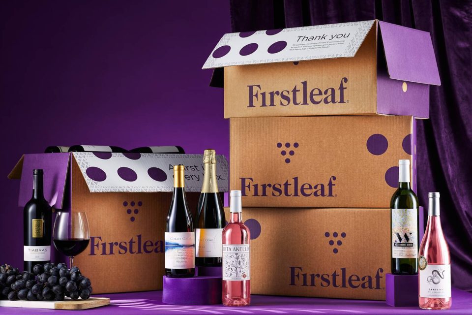 A variety of wine bottles are displayed on a purple surface alongside two large cardboard boxes labeled "Firstleaf." There are also two wine glasses and a bunch of grapes in the foreground. The background features a purple curtain.