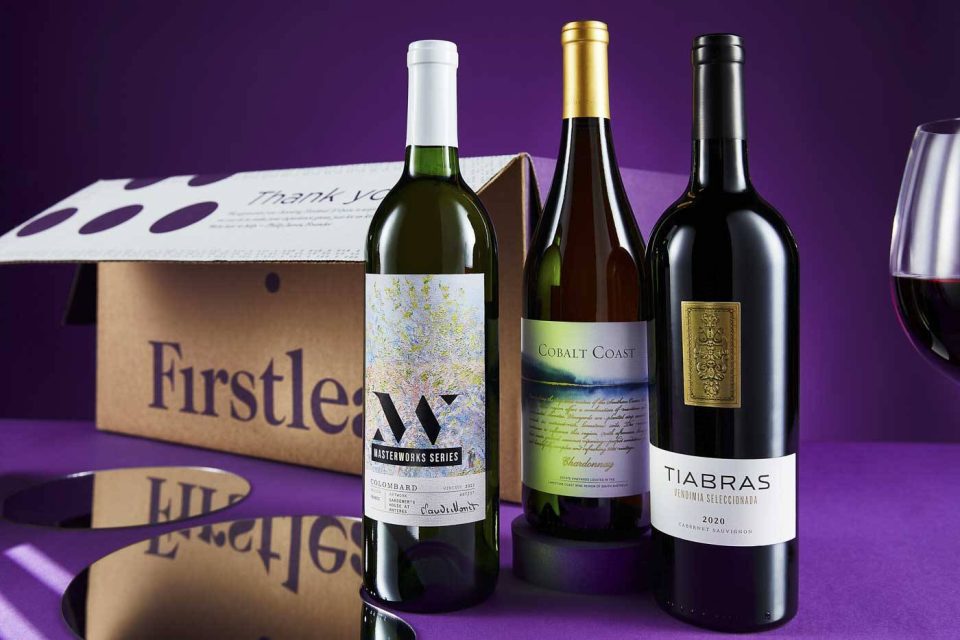 Three wine bottles are displayed against a purple background. They are placed in front of a partially open Firstleaf box. A glass of red wine is visible on the right. The labels feature artistic designs with names: Alloy, Cobalt Coast, and Tiabras.