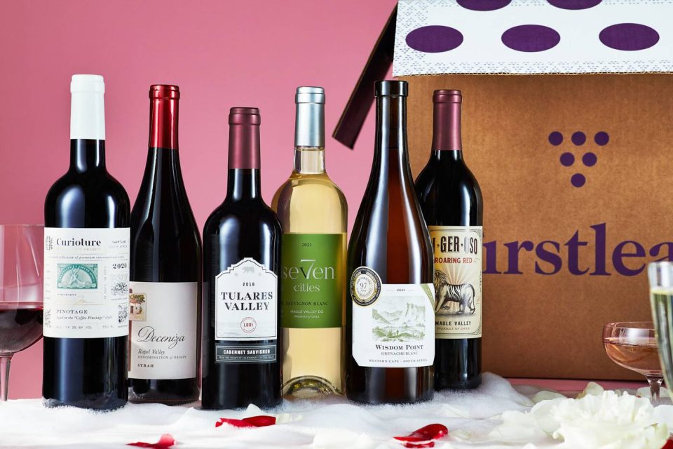 A selection of six wine bottles, including red and white varieties, displayed in front of a Firstleaf box. The background is a soft pink, and there are scattered rose petals and wine glasses around the setup.