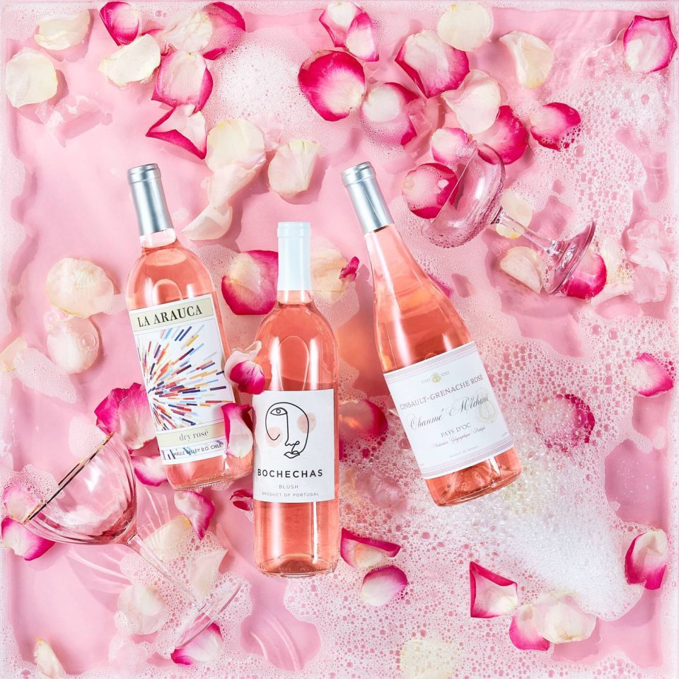 Three bottles of rosé wine on a pink surface scattered with rose petals and surrounded by bubbles. Two empty glasses lie beside the bottles, one upright and the other overturned.