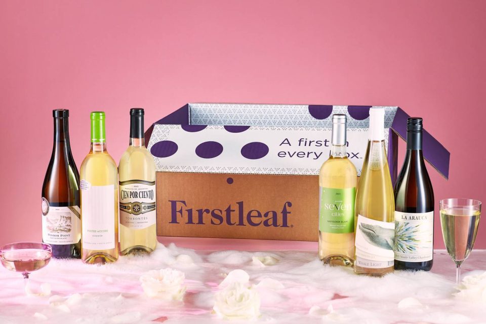 A Firstleaf box with six bottles of assorted white and rosé wines is displayed against a pink background. Two wine glasses, one with white wine, are placed near the box amid soft, white fabric and scattered white flowers.