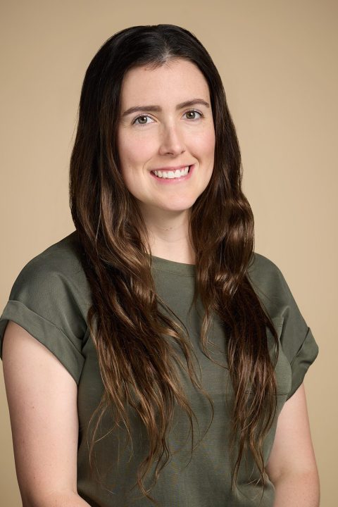 Meet the team: A person with long brown hair and a green shirt smiles warmly against a plain beige background.