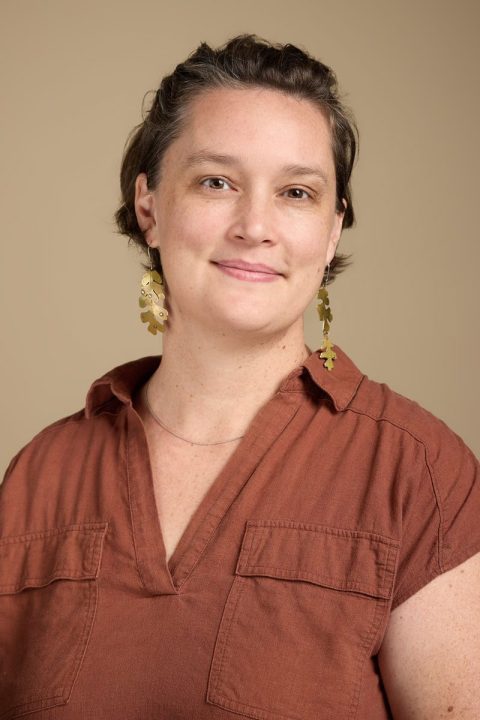 Meet the team member with short hair, wearing a rust-colored shirt and gold leaf-shaped earrings, smiling warmly against a beige background.