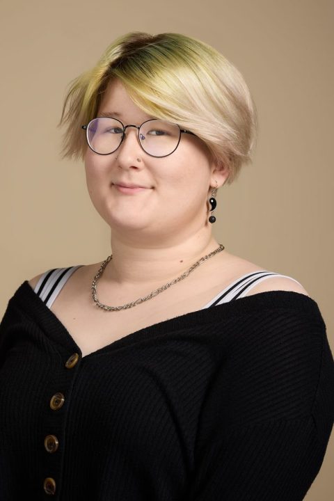 Meet the team member with short blonde and greenish hair, wearing round glasses and a black off-the-shoulder sweater adorned with buttons. Their ensemble is completed with a chain necklace and earrings as they stand against a beige background.