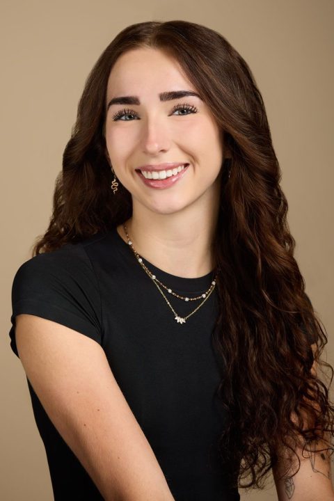 Meet the team: A woman with long, wavy brown hair smiles against a beige background. She is wearing a black short-sleeved top, earrings, and layered necklaces.