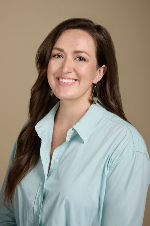 Meet the team: A person with long brown hair is smiling in front of a neutral background, wearing a light blue shirt and triangle-shaped earrings.