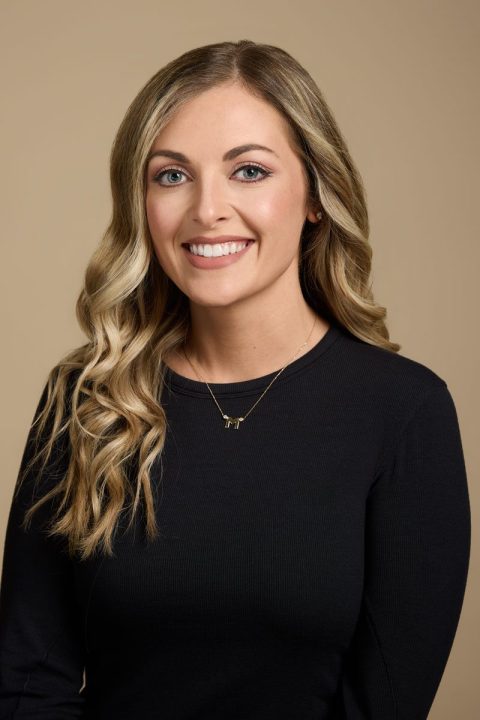 Meet the team: A woman with long, wavy blonde hair smiles at the camera. She is wearing a black top and a gold necklace against a neutral beige background.