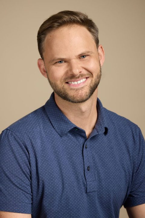 Meet the team: A person with short brown hair and a beard is smiling. They are dressed in a blue collared shirt with small white dots against a neutral backdrop, embodying friendly professionalism.