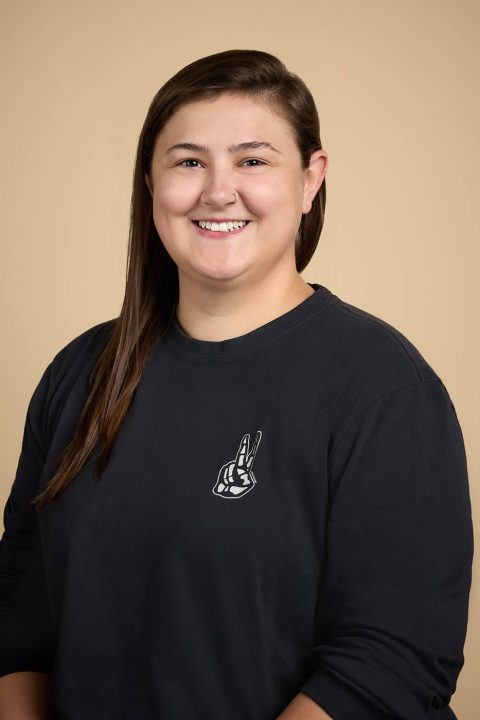 Meet the team: A person with long brown hair smiles warmly, wearing a black sweatshirt adorned with a white graphic of a hand making a peace sign. The backdrop is a plain beige color, complementing their friendly demeanor.