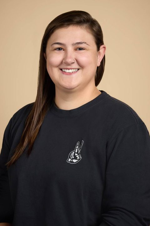 Meet the team: A person with long brown hair smiles warmly, wearing a black sweatshirt adorned with a white graphic of a hand making a peace sign. The backdrop is a plain beige color, complementing their friendly demeanor.