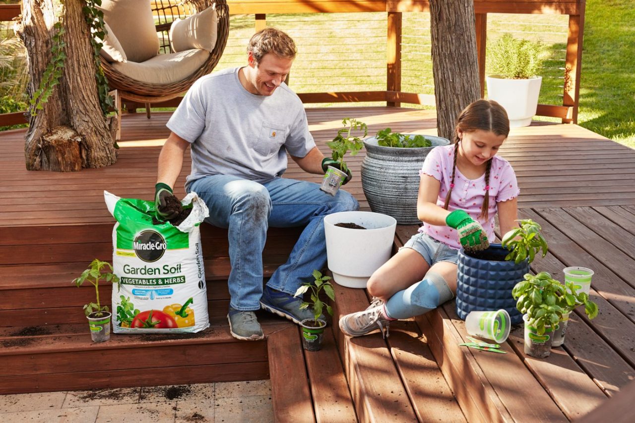 A man and a young girl are gardening on a wooden deck. They're planting small plants into pots using garden soil. The girl is smiling, and they're both wearing gloves. A hammock and green lawn are visible in the background.