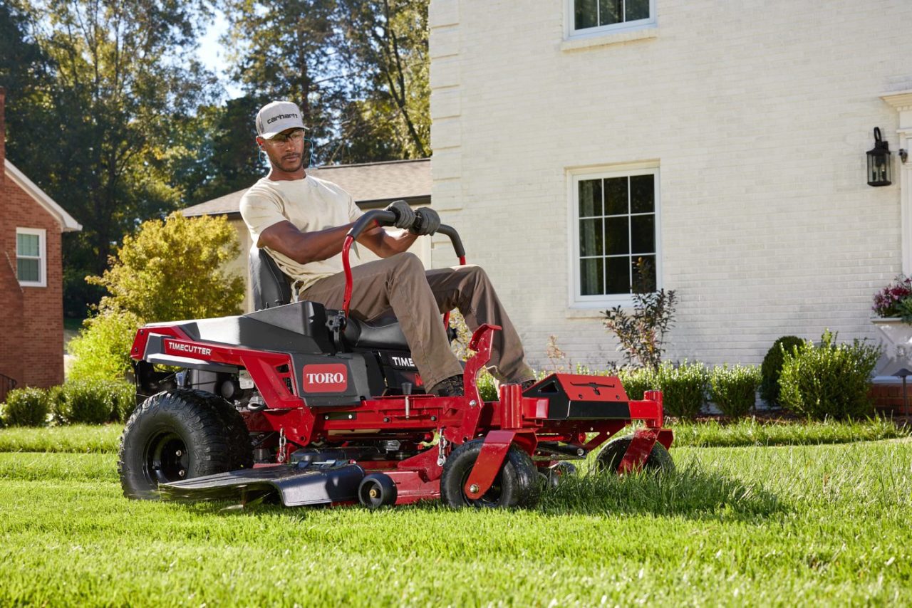 A person is mowing a lawn using a red Toro riding lawnmower. They are wearing a grey cap and sunglasses. In the background is a white brick house, green bushes, and trees.