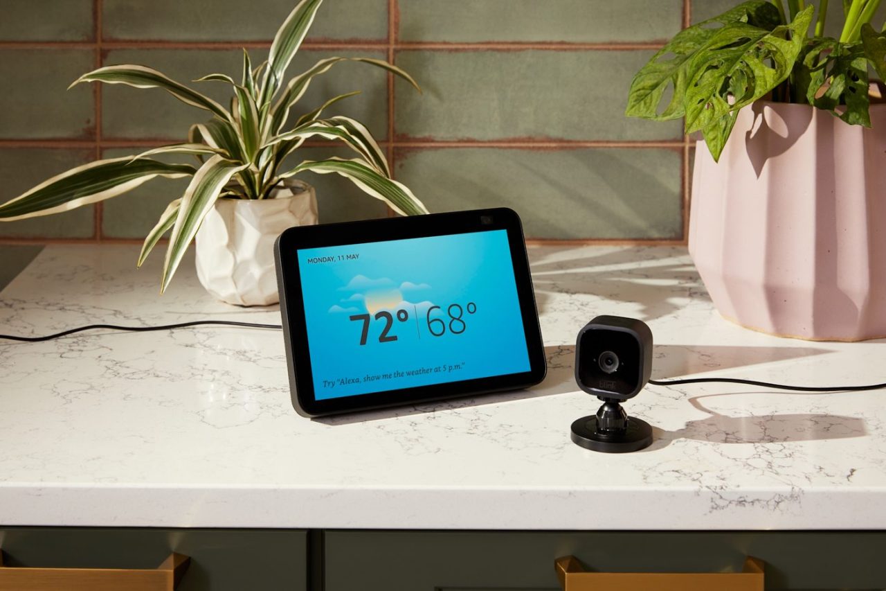 A smart display on a kitchen counter shows the weather as 72°F and 68°F with a cloud graphic. A black security camera is nearby. Two potted plants sit on the counter, and there's a green tiled backsplash.