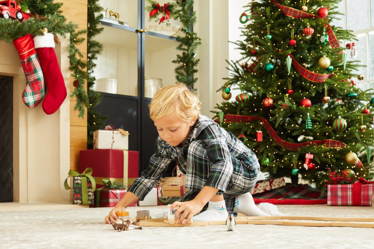 A young child plays with wooden trains on a carpet in a festive living room. A decorated Christmas tree with ornaments and wrapped gifts is in the background, along with a fireplace adorned with stockings.