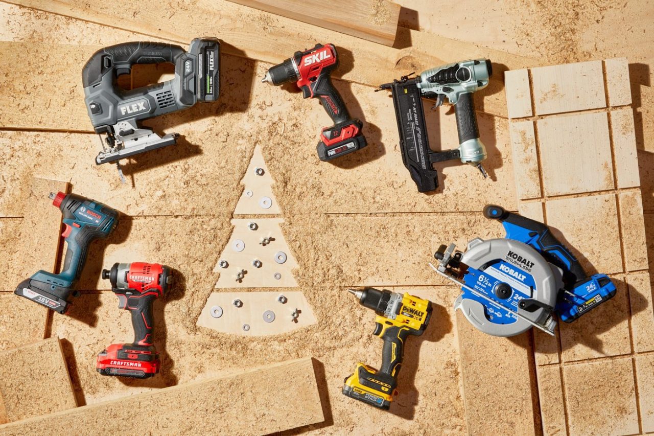 A variety of power tools, including drills, jigsaws, and circular saws, are arranged around wood cutouts forming a Christmas tree shape on a sawdust-covered surface.