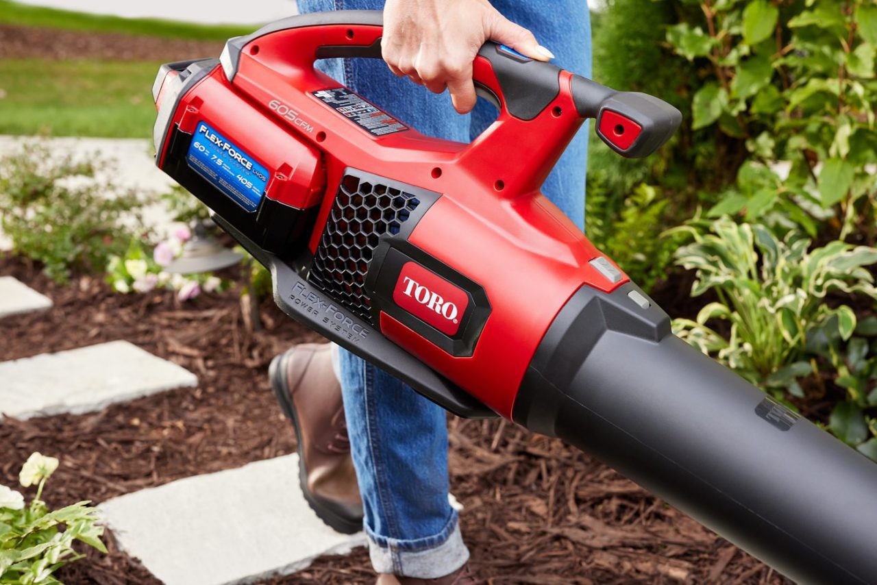 A person holding a red Toro leaf blower outdoors near a garden path. The device is angled downwards, showing a battery pack labeled "60V Max" and "Flex-Force Power System." The ground is covered with mulch and small plants.