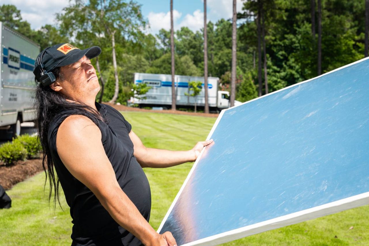 A person with long hair and a cap is outdoors, holding a large reflective panel under bright sunlight. There are trees and parked trucks in the background, with a grassy area surrounding them.